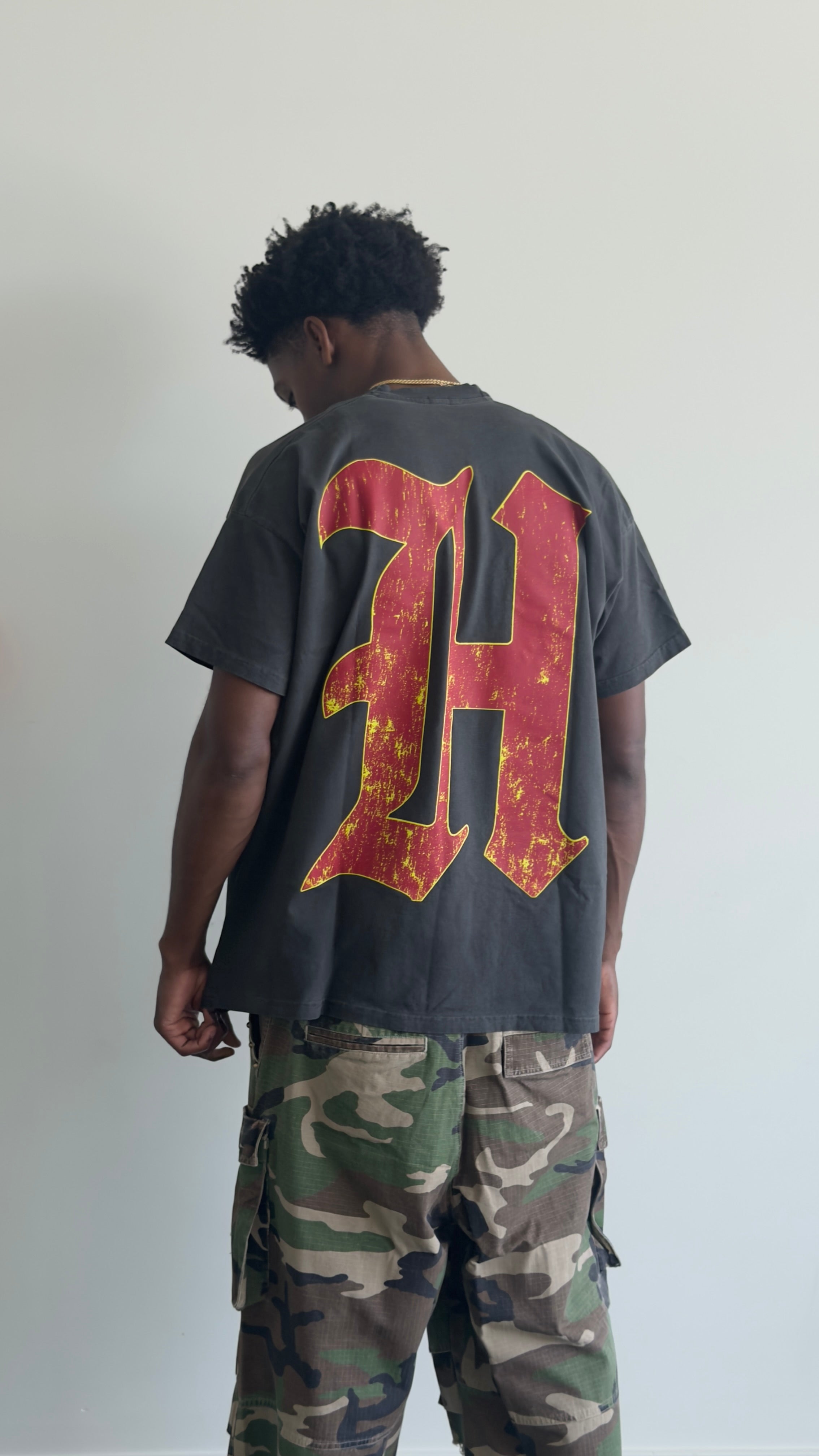 A man viewed from behind wearing a faded black t-shirt with a large distressed red and yellow letter 'H' on the back, paired with green camouflage cargo pants, against a plain light gray wall.