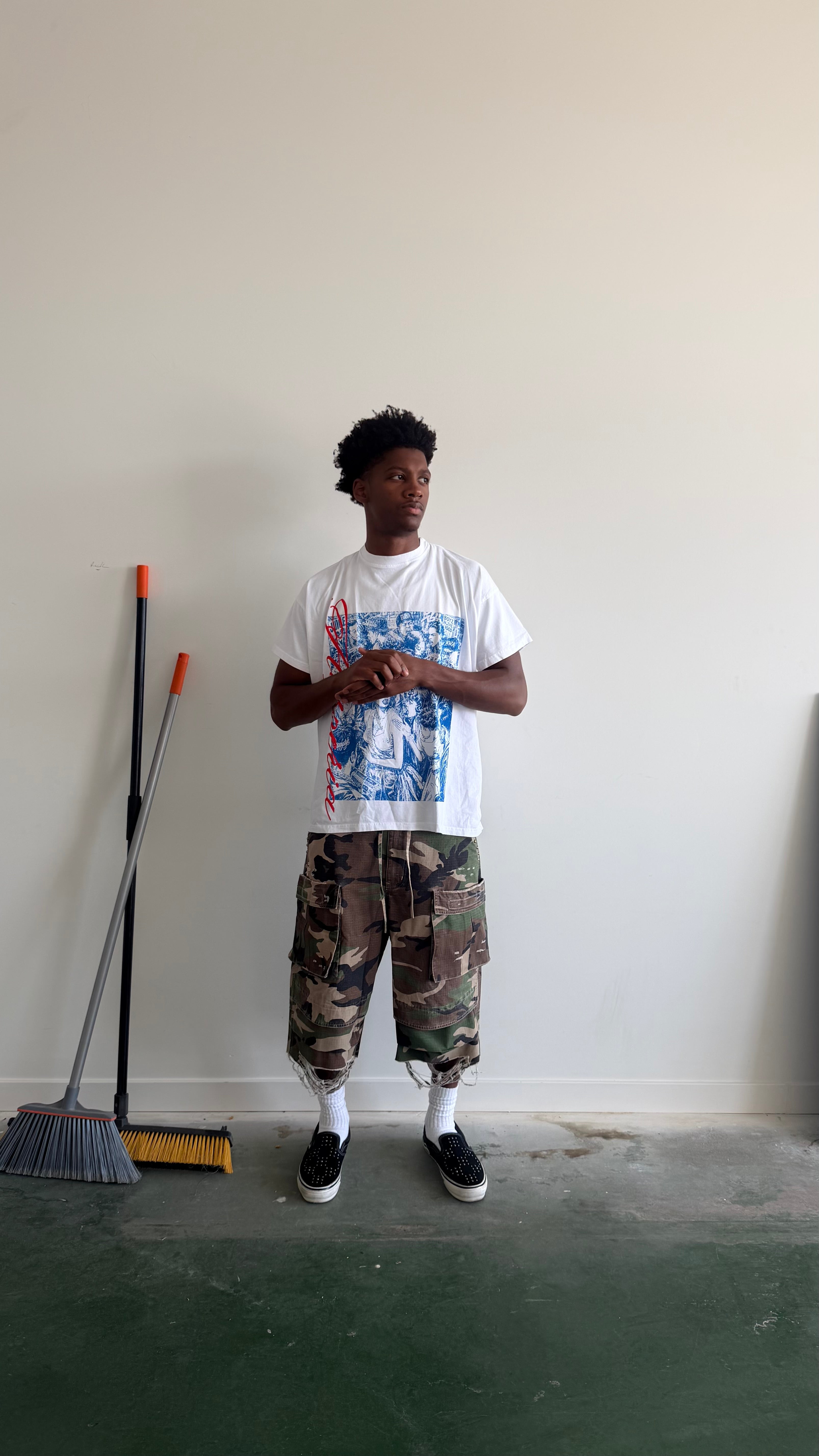 A person stands in a room wearing a white graphic T-shirt, camouflage cargo pants, white socks, and black sneakers. A broom is leaning against the wall on the left.