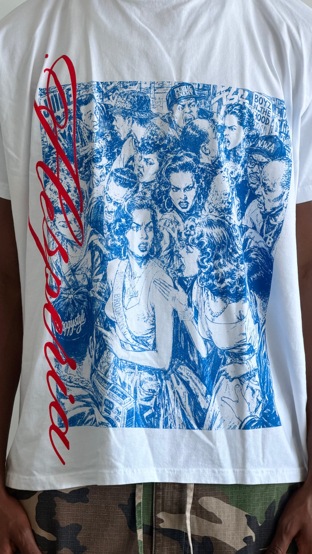 A white graphic t-shirt featuring a large blue print of the band Slipknot on the front, with a red script logo on the left side. The shirt is shown on a person wearing camouflage pants.