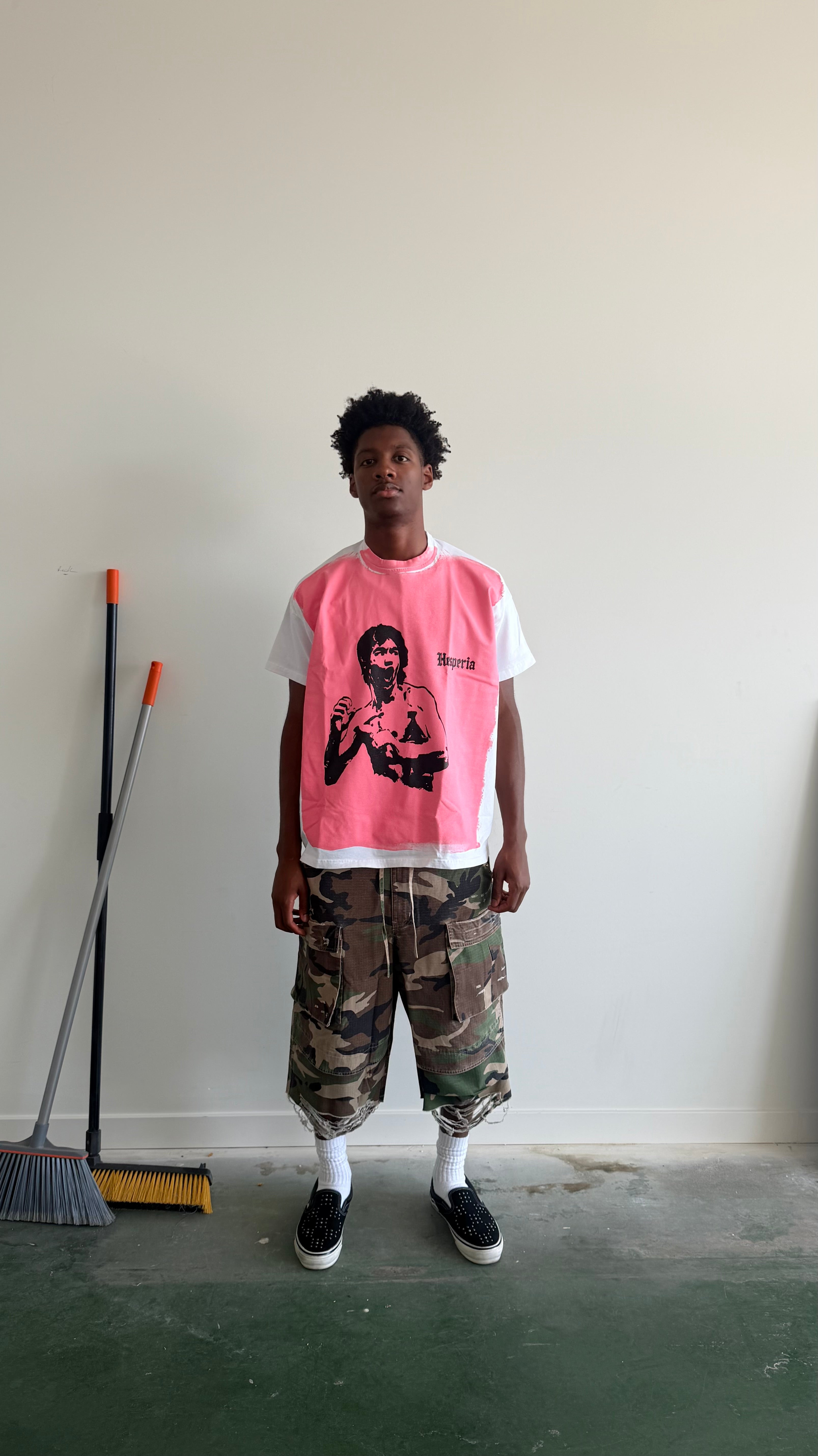 A person stands in a room wearing a pink and white graphic t-shirt, camouflage cargo shorts, white socks, and black sneakers. A broom is leaning against the wall on the left.