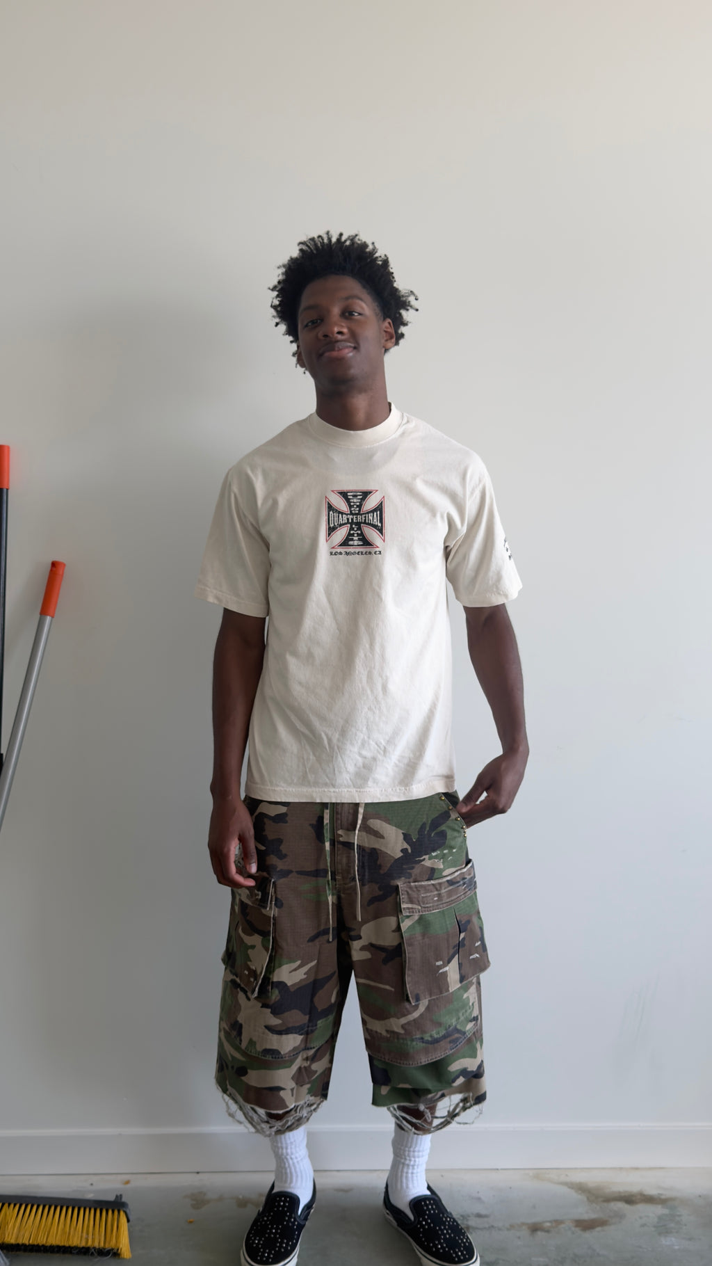 A man wearing a cream-colored t-shirt with a black Iron Cross graphic, paired with green and brown camouflage cargo shorts, white socks, and black slip-on shoes, standing against a plain white wall.