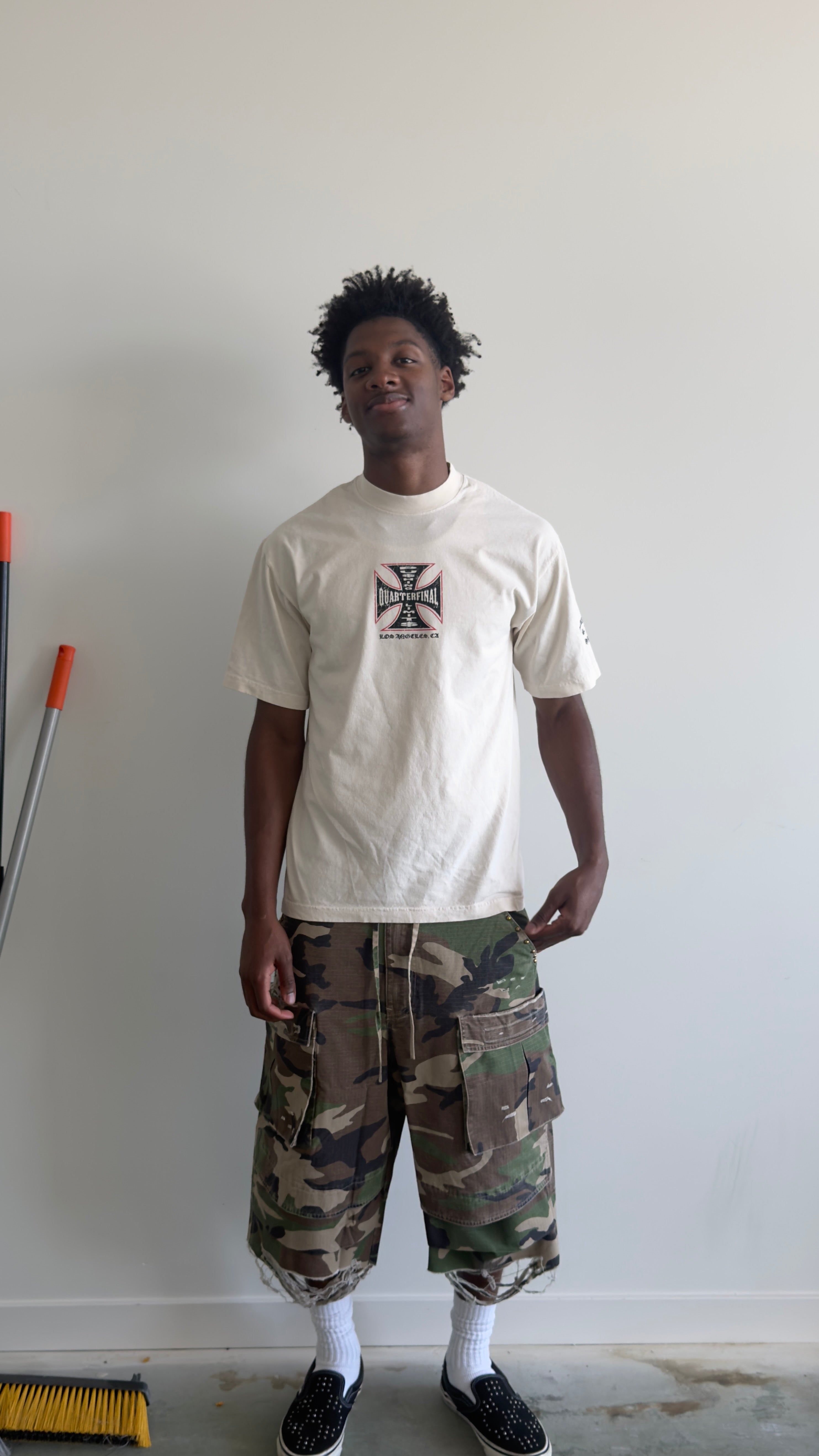 A man wearing a cream-colored t-shirt with a black Iron Cross graphic, paired with green and brown camouflage cargo shorts, white socks, and black slip-on shoes, standing against a plain white wall.
