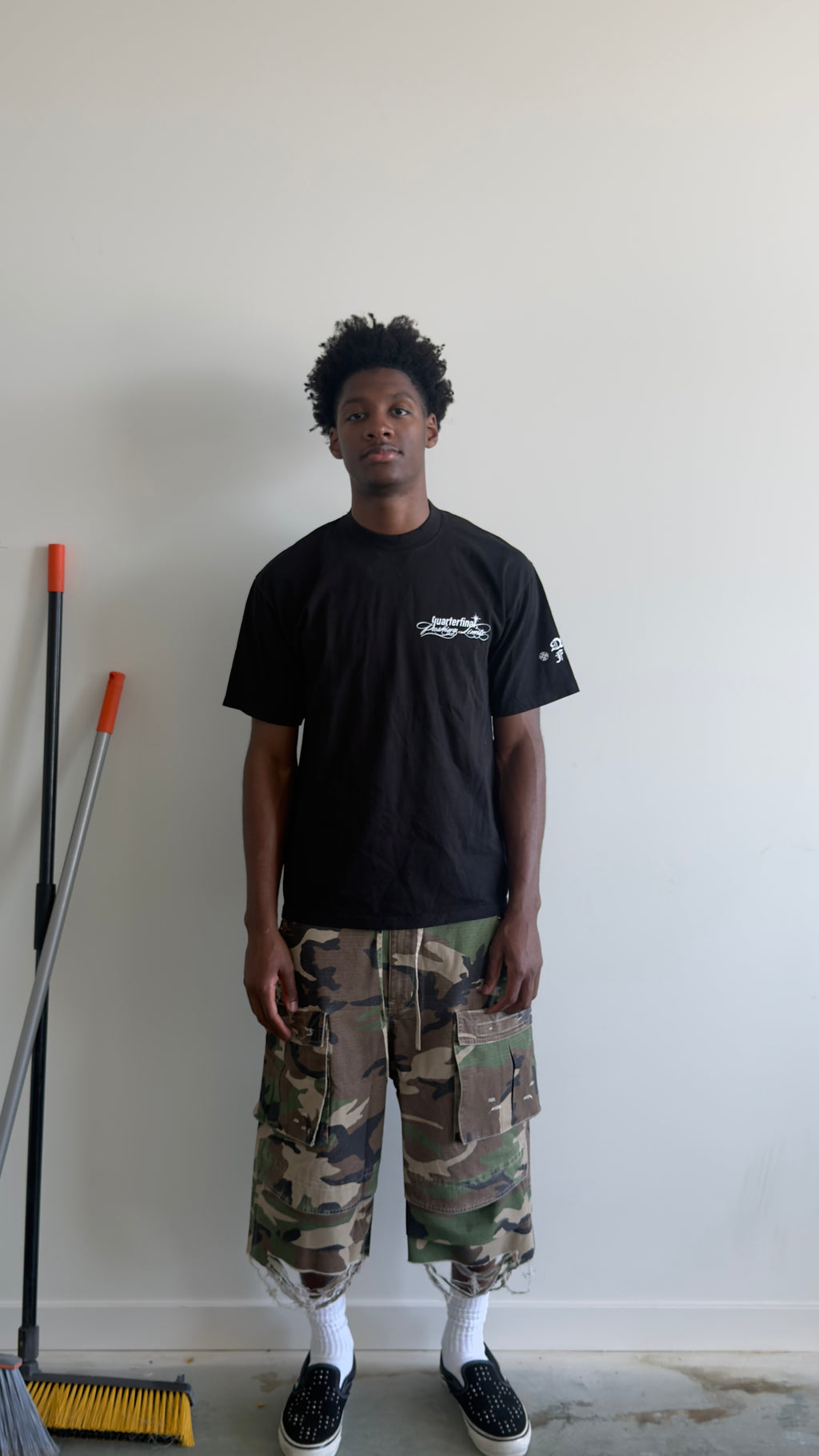 A man stands against a plain white wall wearing a black t-shirt, camouflage cargo shorts, white socks, and black slip-on shoes. A yellow and black broom is leaning against the wall on the left.