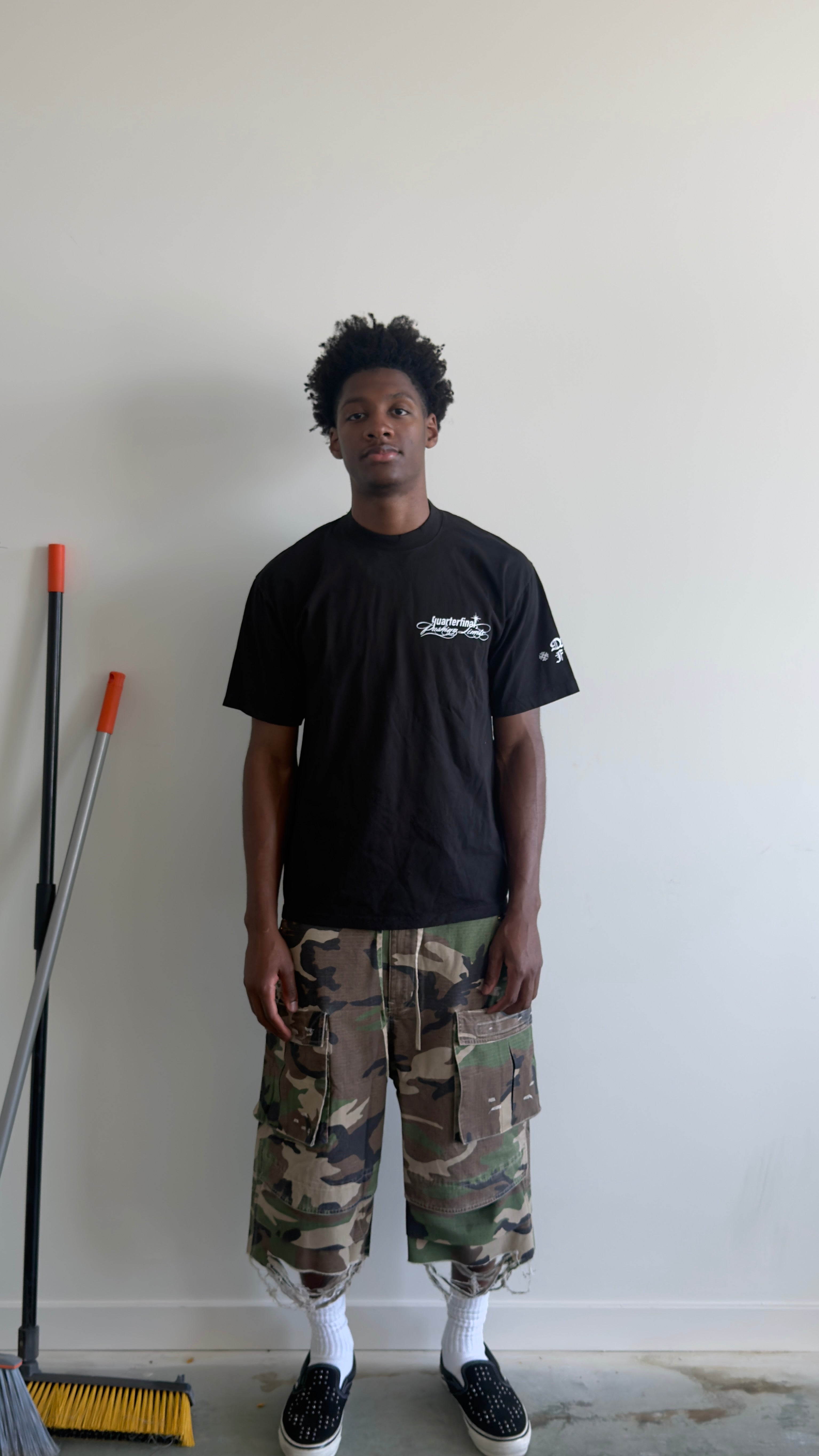 A man stands against a plain white wall wearing a black t-shirt, camouflage cargo shorts, white socks, and black slip-on shoes. A yellow and black broom is leaning against the wall on the left.