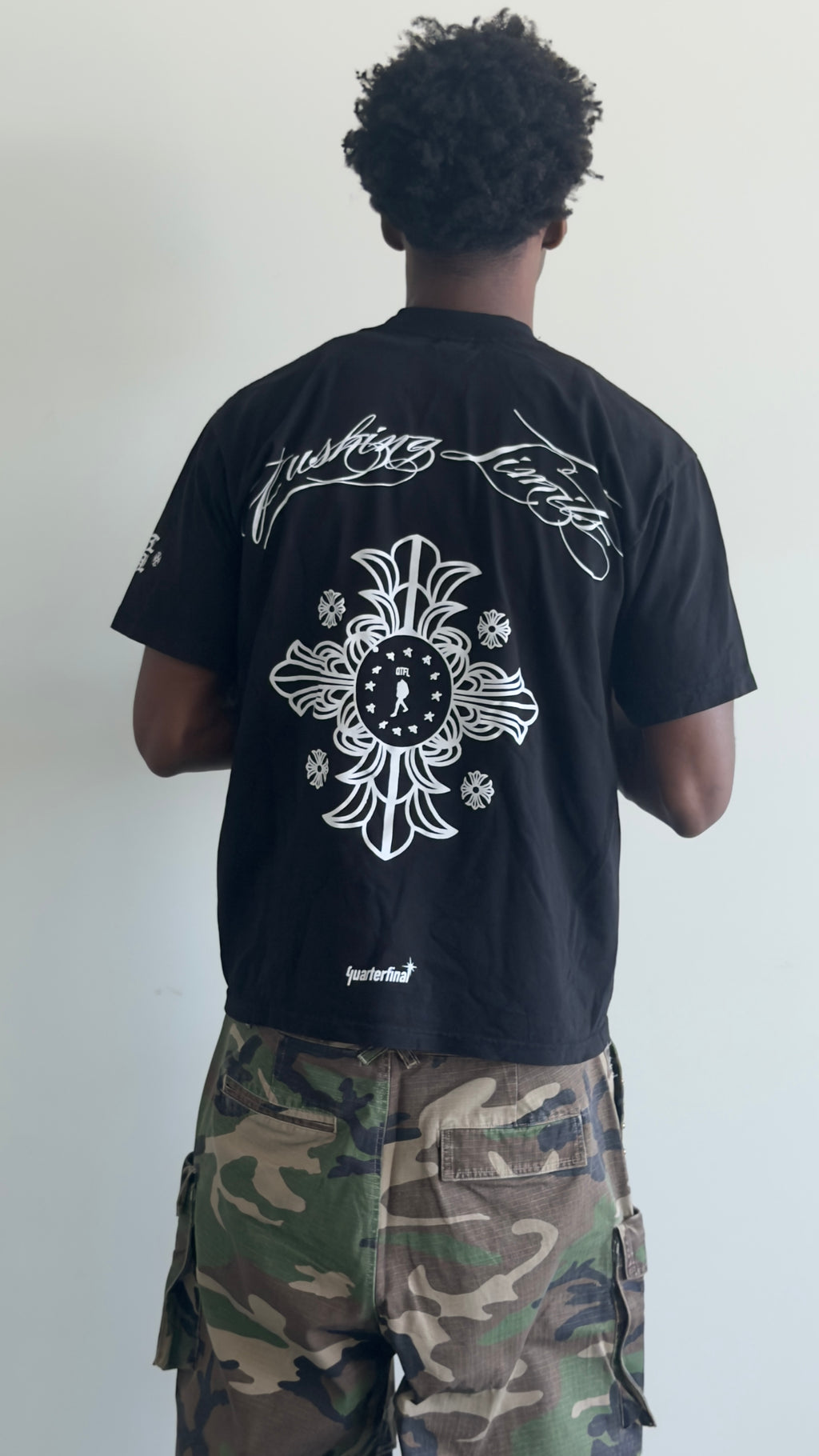 A person shown from the back wearing a black short-sleeve T-shirt with a large white graphic print on the back and camouflage cargo pants, standing against a plain light wall.