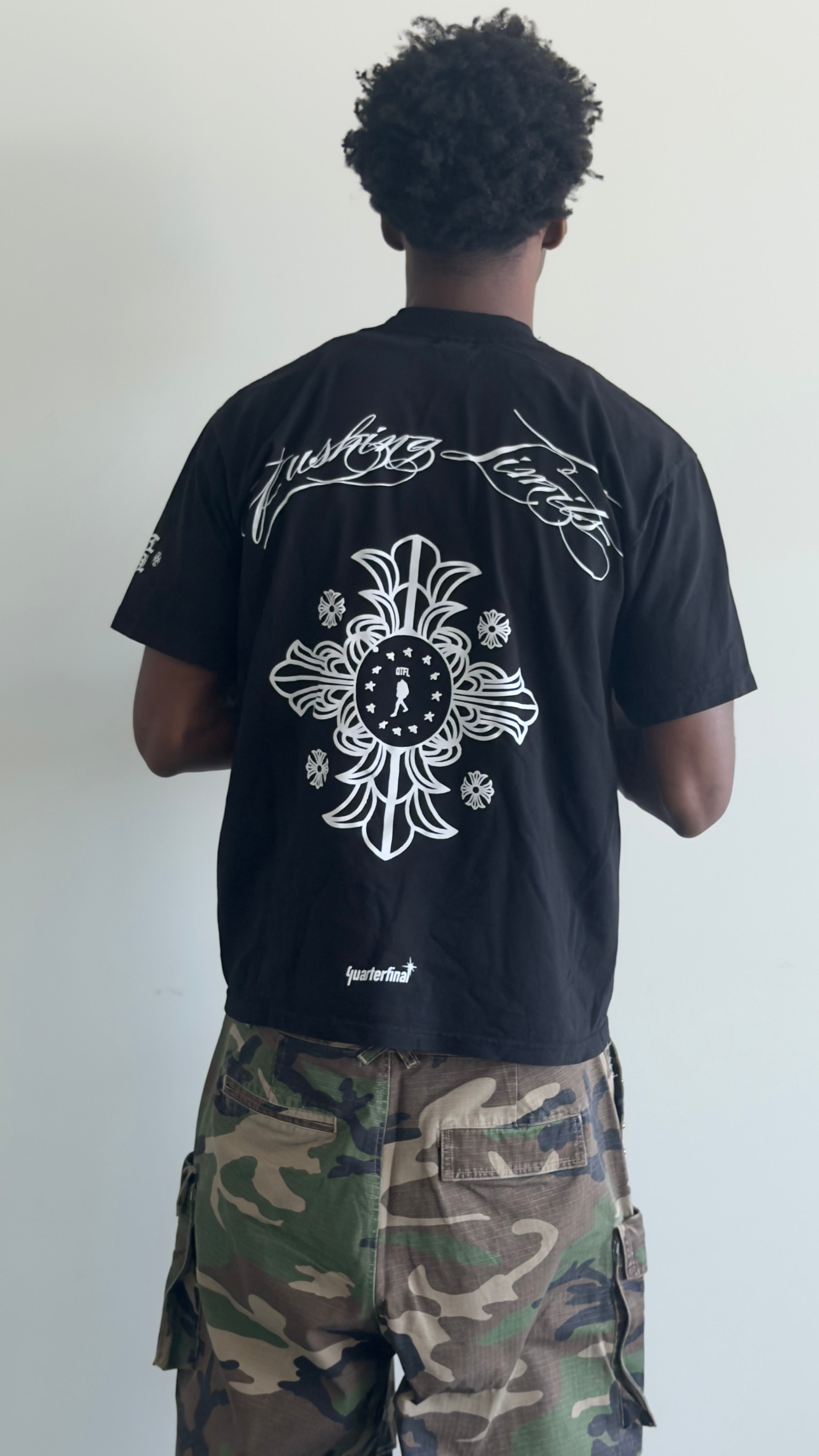 A person shown from the back wearing a black short-sleeve T-shirt with a large white graphic print on the back and camouflage cargo pants, standing against a plain light wall.