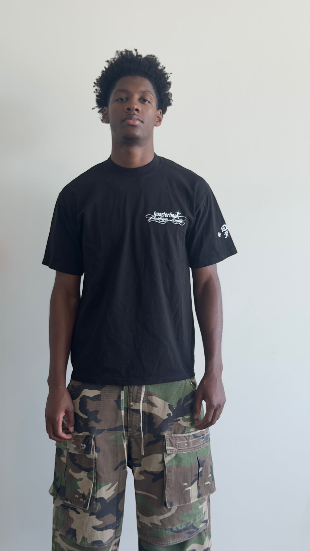 A man wearing a black crewneck t-shirt with white graphic prints on the chest and sleeve, paired with green and brown camouflage cargo pants, standing against a plain off-white wall.