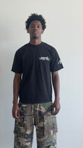 A man wearing a black crewneck t-shirt with white graphic prints on the chest and sleeve, paired with green and brown camouflage cargo pants, standing against a plain off-white wall.