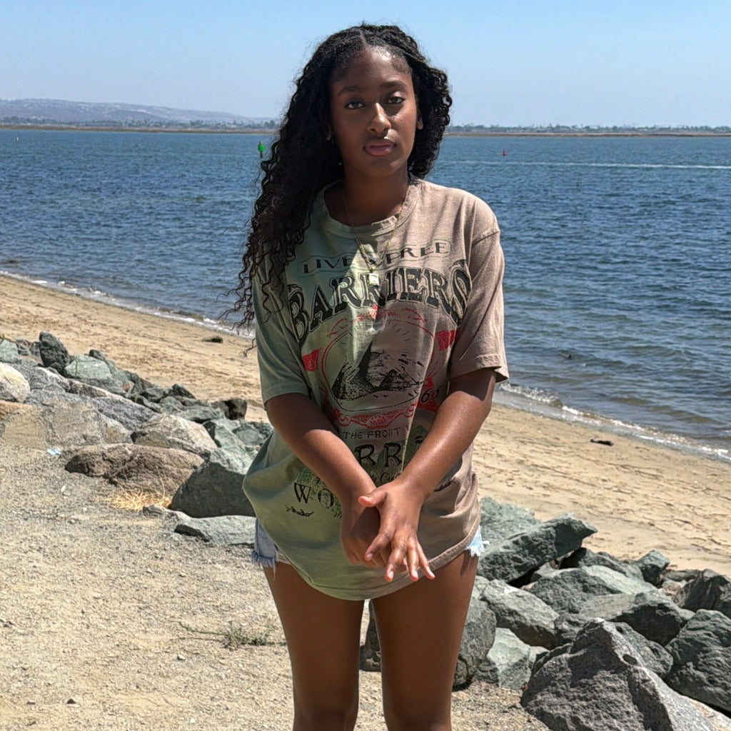 A young woman stands on a sandy shoreline with rocks and a large body of water behind her under a clear blue sky. She is wearing a light beige graphic t-shirt and matching shorts.