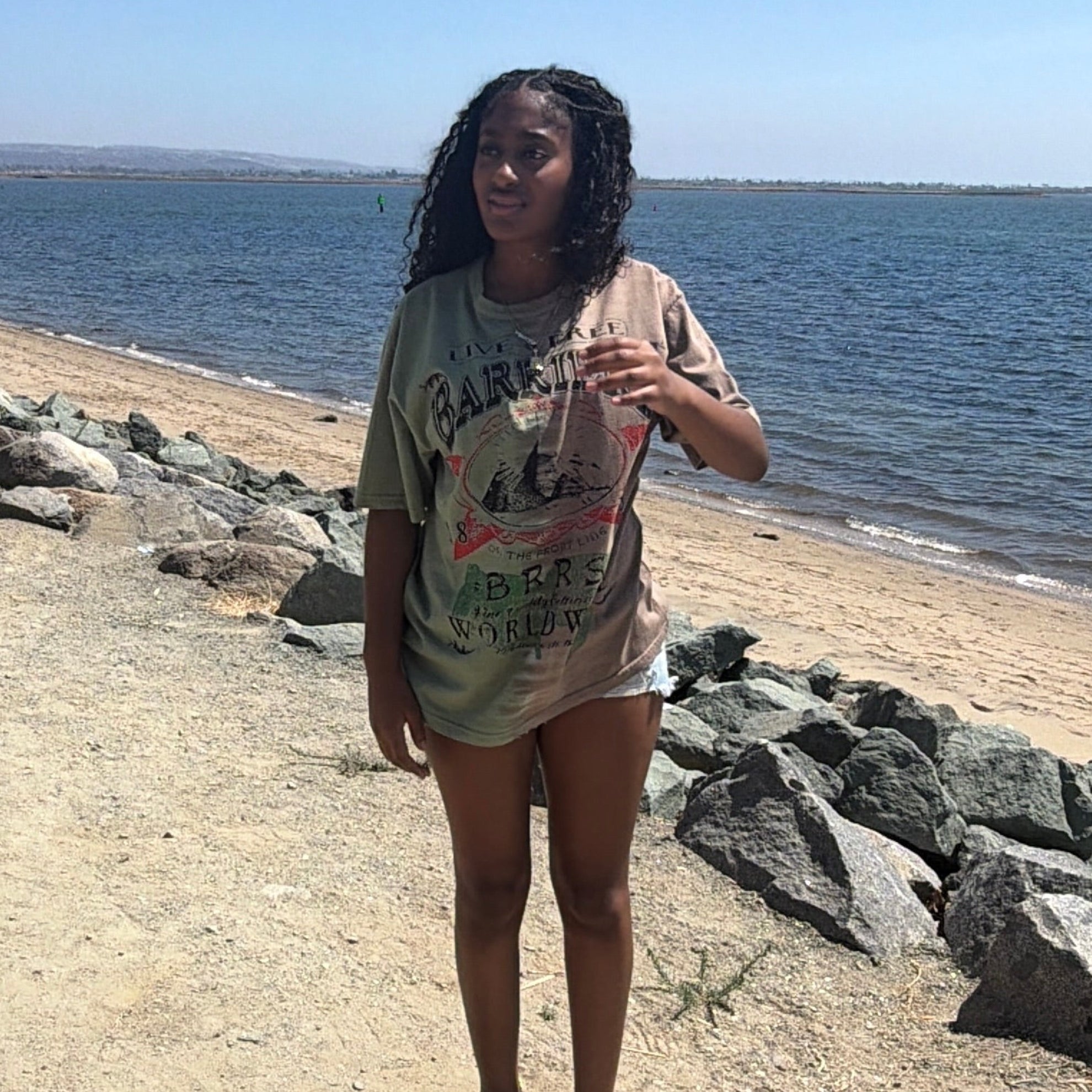 A woman stands on a sandy path next to a rocky shoreline with water and a clear blue sky in the background, wearing a light beige graphic t-shirt and shorts.