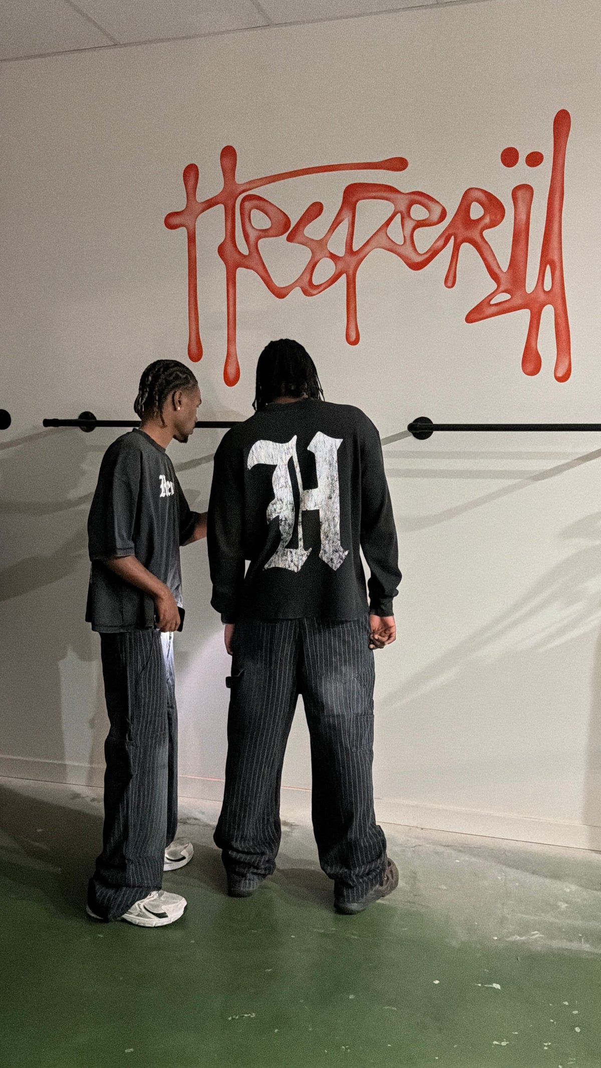Two people standing indoors in front of a white wall with red graffiti-style lettering and a large white 'H' on the back of a black sweatshirt. One person wears a dark gray T-shirt and dark pinstriped pants, and the other wears a black sweatshirt with a large white 'H' on the back and dark pinstriped pants.