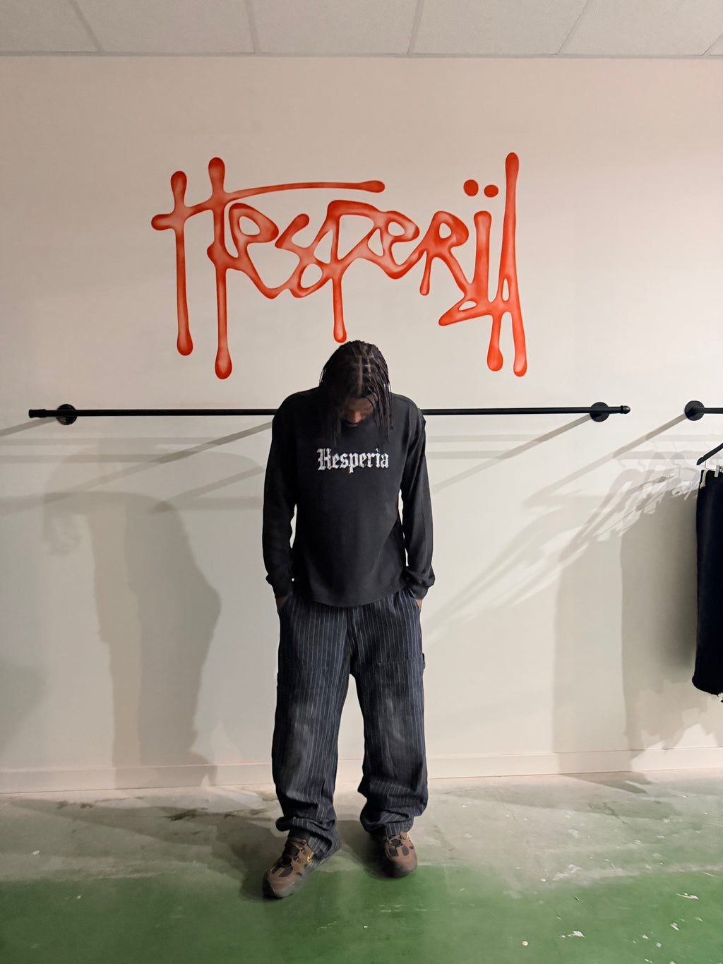 A person stands in a room wearing a black long-sleeve t-shirt with 'Hesperia' printed in white Gothic font, paired with dark gray pinstriped pants. The background features a white wall with a large orange graffiti-style 'Hesperia' logo and a clothing rack.