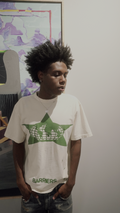 A man with an afro hairstyle wearing a white t-shirt with a green star and globe graphic, standing in front of a light gray wall with a framed art piece in the background.