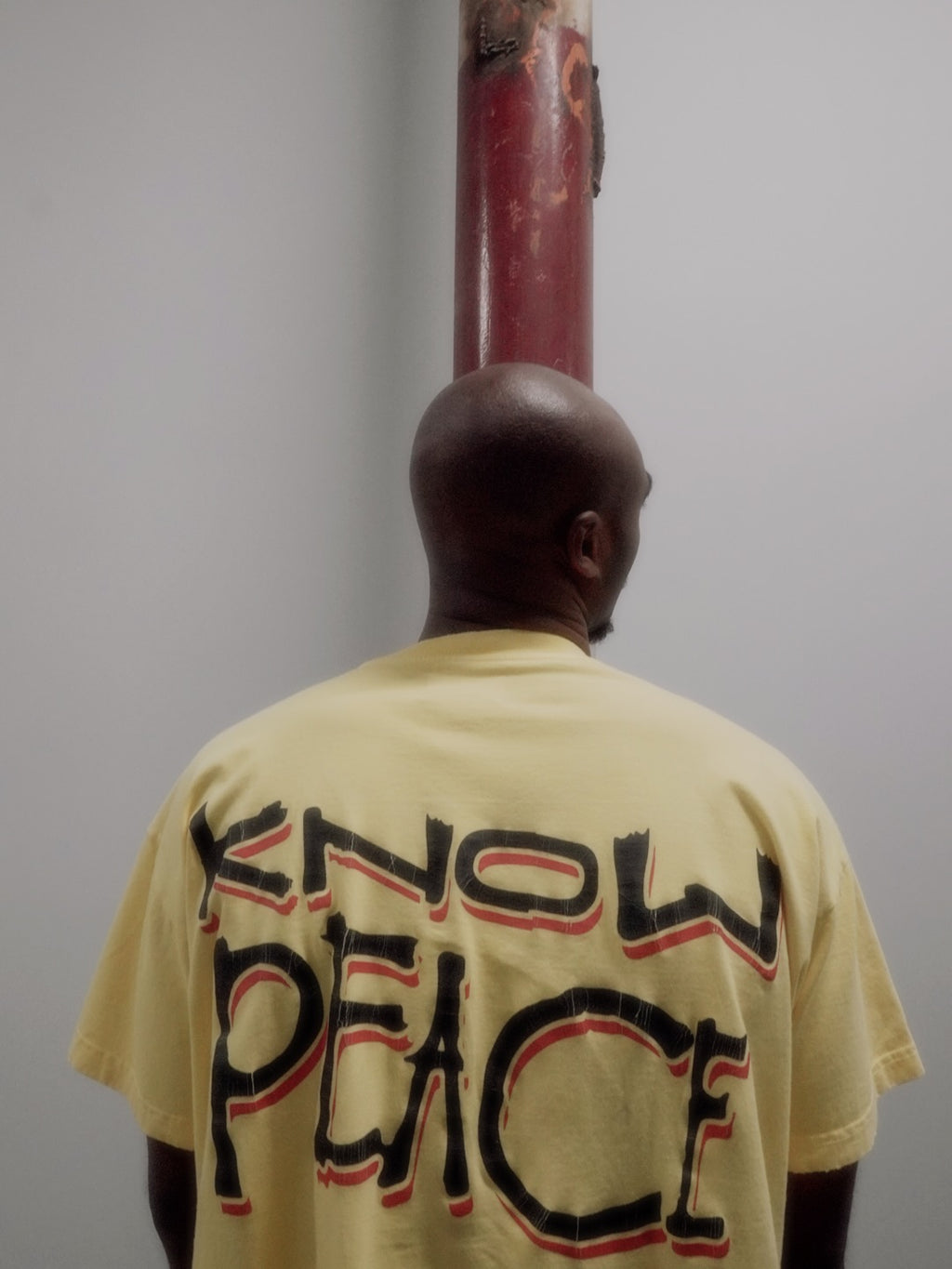 A man viewed from behind wearing a pale yellow t-shirt with 'KNOW PEACE' printed in black and red on the back, standing in front of a red metal pole against a plain gray wall.