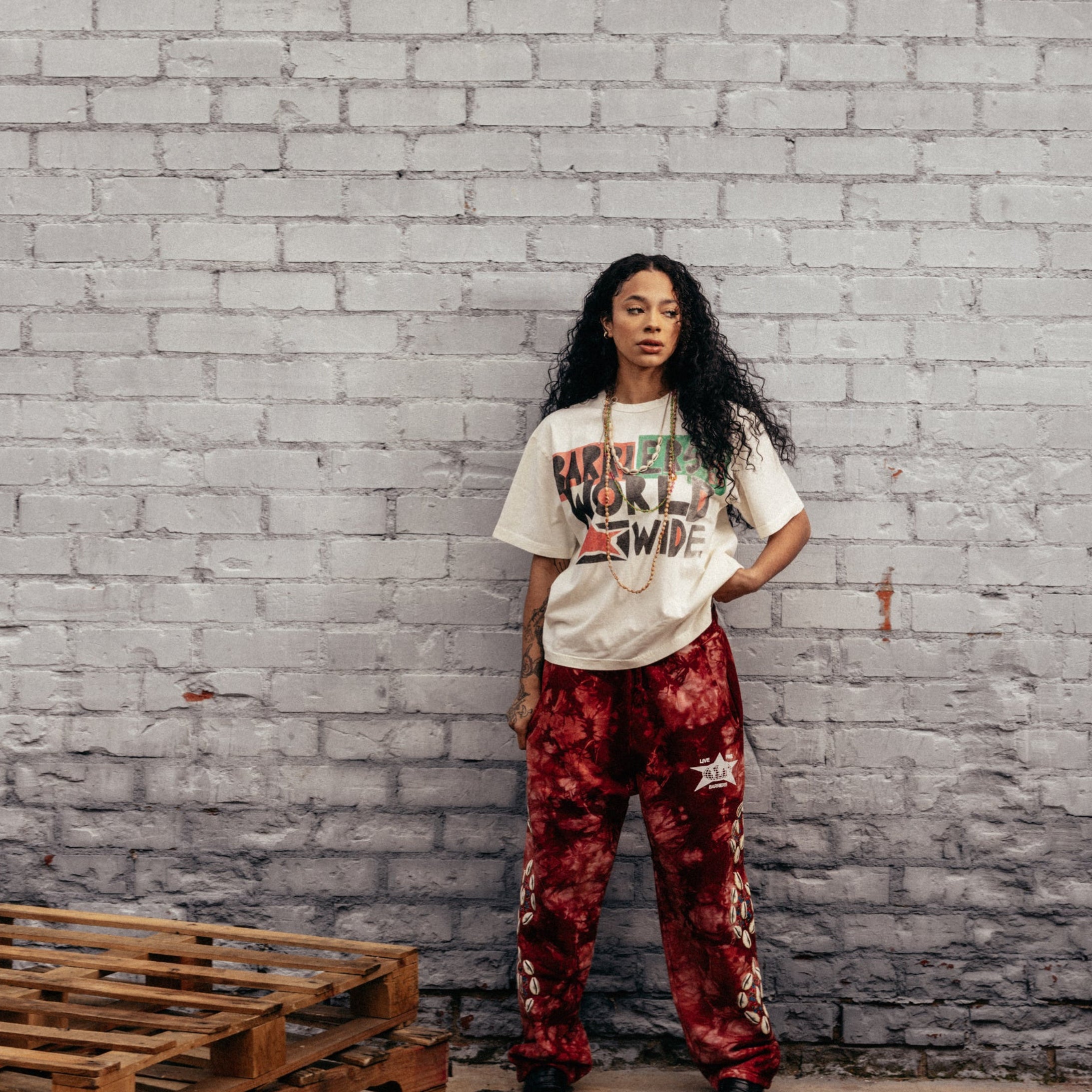 A woman stands against a gray brick wall wearing a white graphic t-shirt and red patterned pants, with wooden pallets on the concrete floor.